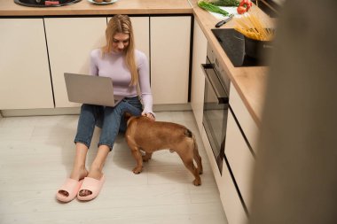 Mutfak zemininde oturmuş, taşınabilir bilgisayarlı gülümseyen kadın köpeğini okşuyor.