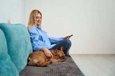 Smiling young female seated on sofa holding remote control unit and stroking her pet