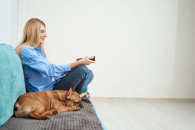 Side view of smiling woman with remote control unit in hand sitting on sofa beside dog
