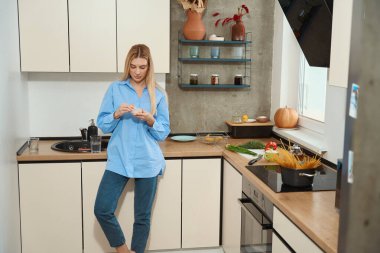 Serious female leaning against kitchen counter while taking capsule out of transparent plastic medicine storage box