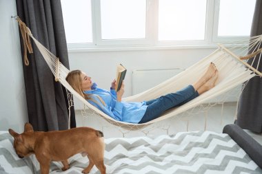Side view of female with book lying in hammock while her dog walking on bed