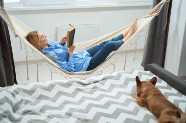 Side view of woman reading book in hammock while her pet sleeping on bed
