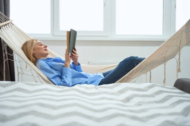 Side view of relaxed young woman with book lying in hammock indoors