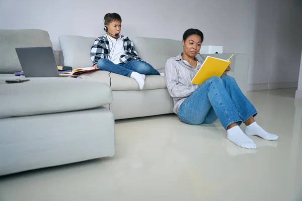 Modern teenager on comfortable home online learning, boy and his mother are located in room with a laptop, headset, books
