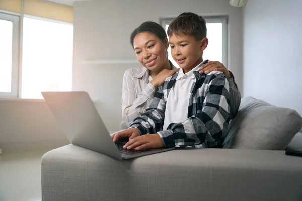 Mom and son are sitting on the sofa with a laptop, a caring woman helps her son in online learning