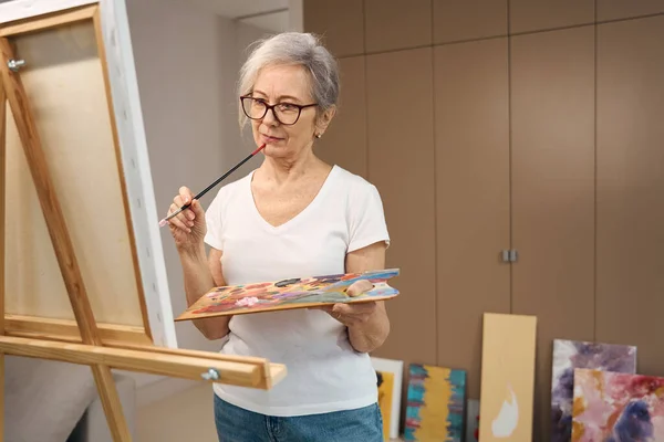 スレンダーなおばあさんが広々とした部屋で絵を描き 彼女の以前の作品はクローゼットのそばにあります — ストック写真