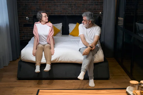 stock image Elderly couple sitting on bed in bedroom, looking at each other, preparing for sleep