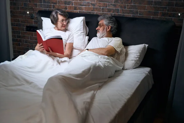 Старая Женщина Мужчина Лежат Кровати Спальне Жена Читает Книгу Мужу — стоковое фото