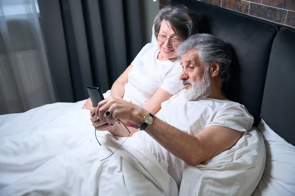 Elderly Couple Lying Bed Bedroom Male Holding Mobile Phone Having — стоковое фото
