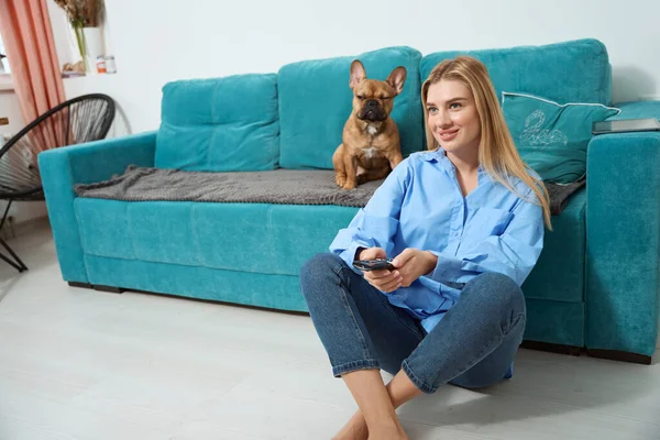 Smiling young woman changing channels on TV in presence of her serious dog
