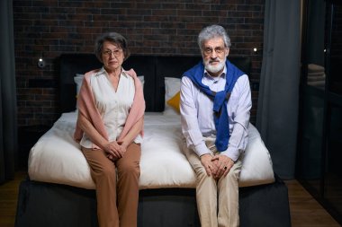 Üzgün sakallı adam ve yoldaşı büyük bir yatakta oturuyorlar, eşler rahat bir otelde kalıyorlar.