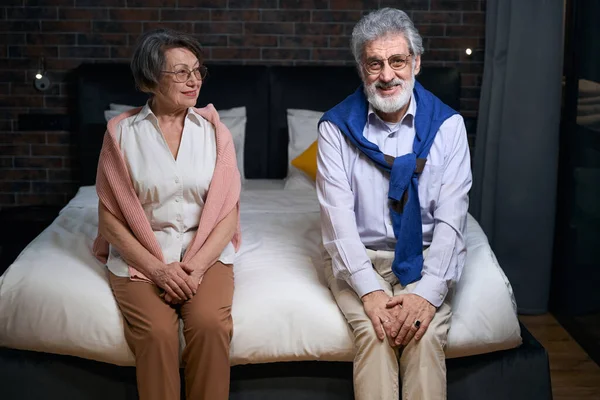 心满意足的白发苍苍的男人和他的伴侣坐在一张大床上 这对夫妇住在一家舒适的旅馆里 — 图库照片