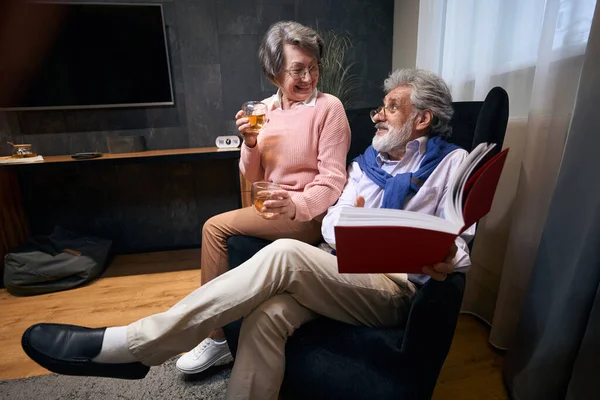 老夫妇舒适地坐在扶手椅上看书喝酒 人们愉快地交流着 — 图库照片