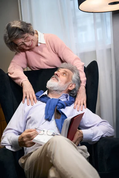 大胡子的祖父坐在舒适的扶手椅上看书 一位老太太抱着他的肩膀 — 图库照片