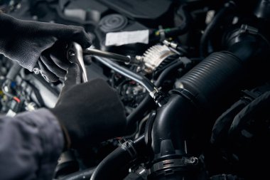 Koruyucu eldivenli bir adam açık kapüşonlu arabanın yanında duruyor ve alternatör kayışının gerginliğini kontrol ediyor.