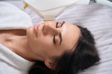 Salondaki kanepede uzanan genç bir kadın koruyucu eldivenli bir sıhhiye yüz için IPL fotorejuvenation yapıyor.