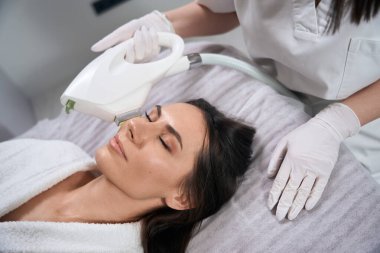 Salondaki kanepede uzanan kadın, koruyucu eldivenli doktor yüzü için IPL fotorejuvenation yapıyor.