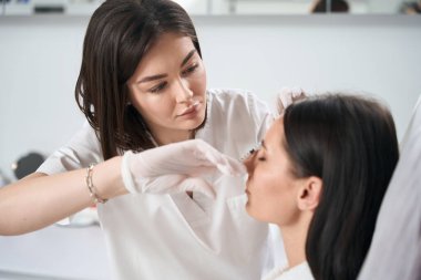 Güzeller güzeli kadın salonda kanepede oturuyor, koruyucu eldivenli kozmetolog yaşlanma önleyici iğneler yapıyor.