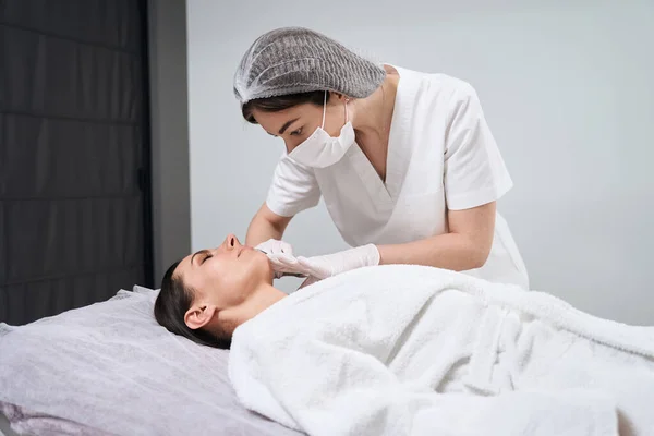 Beautiful lady lying on couch, cosmetologist in protective gloves and mask doing anti-aging massage