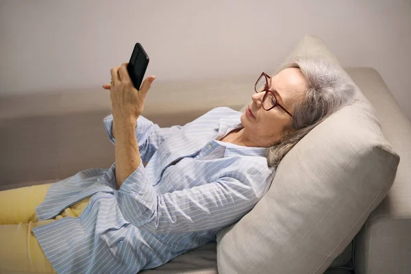 老年妇女在家休息 看着手机里的东西 坐在柔软的沙发上 — 图库照片