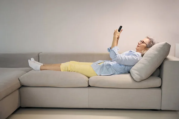 在舒适的家庭环境中 灰白头发的女士躺在沙发上 她用电话 — 图库照片