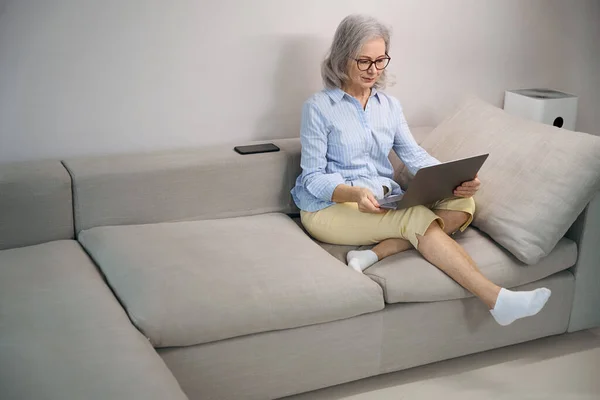 電話の横にある自宅でソファの上にノートパソコンで快適に定住した年金受給者 — ストック写真