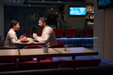 Elinde kahve olan bir erkek, masada salata yiyen bayan arkadaşına bakıyor.