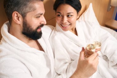 Gülümseyen genç kadın ve mutlu arkadaşı yatakta şampanya bardaklarını tokuşturuyor.