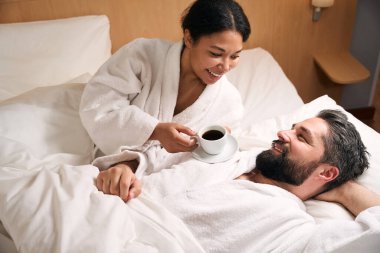Elinde bir fincan kahveyle gülümseyen kadın yatakta mutlu erkek arkadaşının yanında yatıyor.