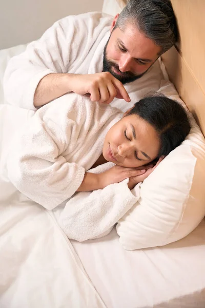 一个爱开玩笑的男人躺在床上 用食指触摸他的女伴的耳朵 — 图库照片