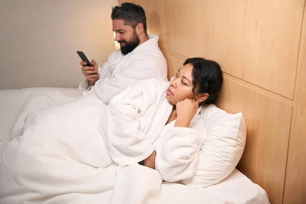 年轻的女性看到她快乐的伴侣在床上用智能手机发短信时 很不高兴 — 图库照片