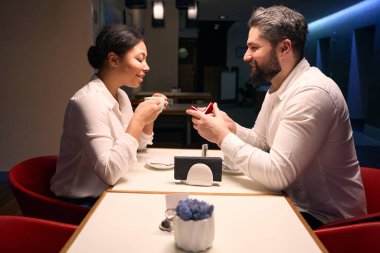 Mutlu genç adam, kafe masasındaki mutlu bayan arkadaşına nişan yüzüğünü takdim ediyor.