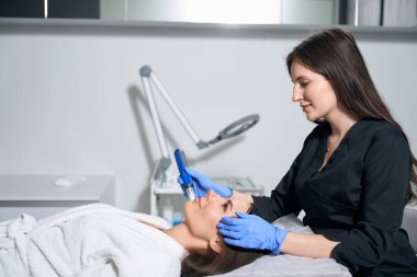 Koruyucu eldivenler ve palto tutma cihazı takan ve kuaförde kadınlara akne tedavisi uygulayan bir tıp çalışanı.