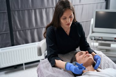 Koruyucu eldivenli ve palto tutan bir kadın kuaförde sivilce tedavisi görüyor.