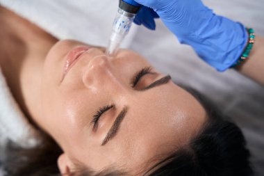 Medical worker in protective gloves holding device and doing post acne treatment to beautiful female in salon clipart