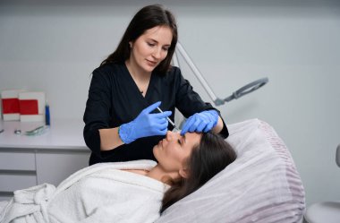 Koruyucu eldivenler ve paltolu bir doktor elinde şırıngayla müşteriye güzellik iğnesi yapıyor.