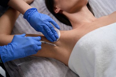 Koruyucu eldivenli ve ceketli kadın şırınga tutuyor ve müşteriye koltukaltı iğnesi yapıyor.