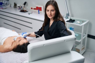 Koruyucu eldivenleri ve ceketi olan kozmetolog kadının yanında oturuyor ve IPL fotorejuvenasyon yapıyor.