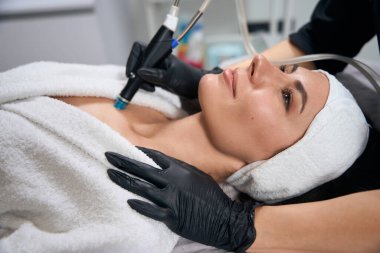 Koruyucu eldivenli kozmetik uzmanı yetişkin bir kadının yanında oturuyor ve ofiste hidroseksüel işlem yapıyor.