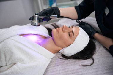 Koruyucu eldivenli doktor yetişkin bir kadının yanında oturuyor ve ofiste terapi prosedürü uyguluyor.