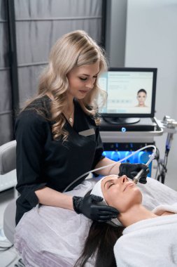 Cosmetologist in protective gloves sitting near pretty woman and doing electroporation procedure for face in salon clipart
