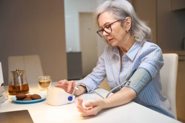 Endişeli yaşlı bayan kan basıncını bir tonometreyle ölçüyor, evde tek başına mutfakta oturuyor.