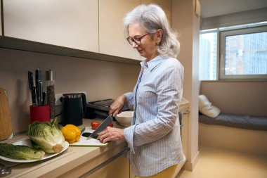 Evdeki mutfakta yaşlı bir kadın salata için sebze kesiyor, boş vakitlerinde yemek pişirmekle meşgul.