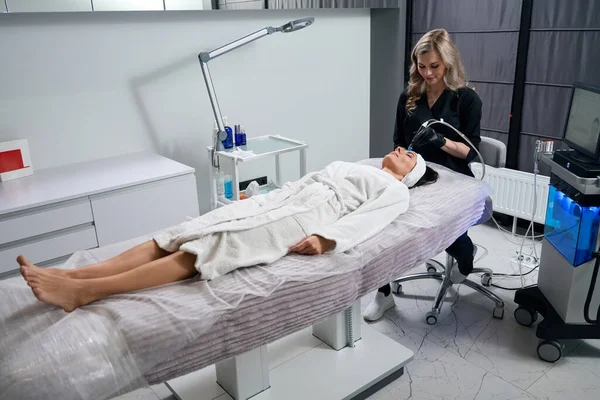 stock image Cosmetologist in protective gloves sitting near beautiful client and doing HydraFacial procedure in salon