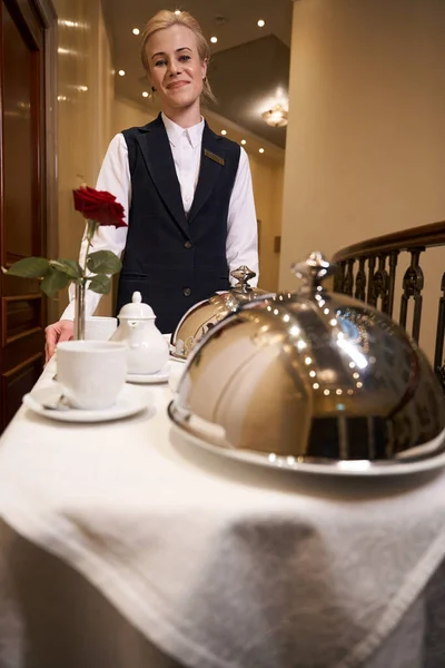 Smiling young woman on cart carrying hot food down hotel corridor, uniformed waitress
