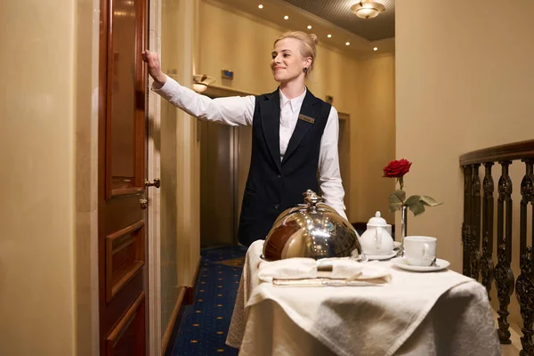Pleasant waitress knocking on the door of the hotel room, she delivered food for a romantic dinner