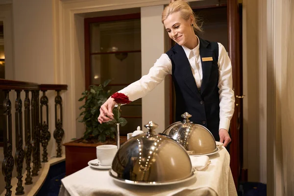 Caring waitress in uniform delivers food to hotel room, everything for romantic meal is on serving table