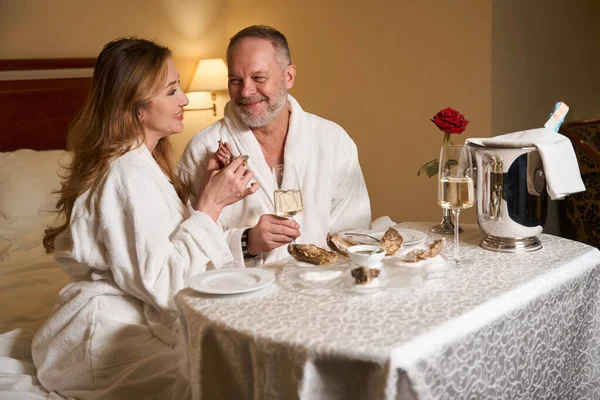 豪華なホテルの部屋でカキとシャンパンで美しいカップルのチャット 彼らは大きなベッドの上に位置しています — ストック写真