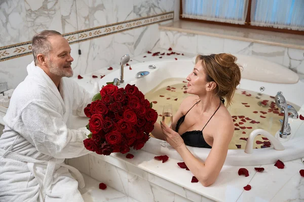 가운을 미소짓는 남자가 아름다운 아내에게 꽃다발을 여인은 욕조에서 장미꽃 잎으로 — 스톡 사진