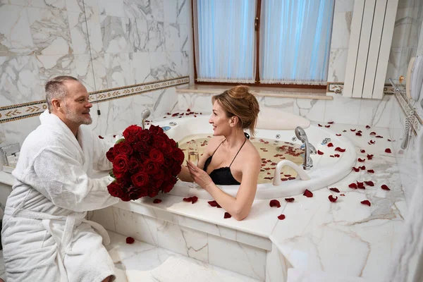 バスローブの髭の男は彼の美しい妻に花束を与えます 女性はシャンパンのガラスでバスルームに飛び出します — ストック写真